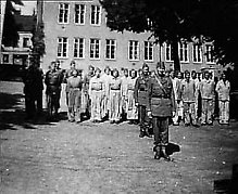 Svart och vit bild med personer som står på rad framför ett ljust tegelhus. Personerna är klädda i uniformer och hatt. 