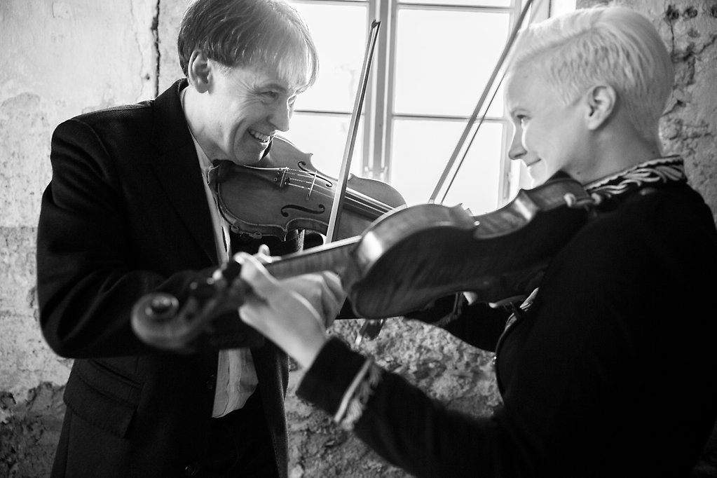 Två spelmän, en man och en kvinna står vända mot varandra och spelar på fiol.