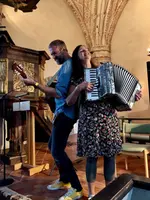 Bild på en man som spelar gitarr och en kvinna som spelar dragspel i en kyrka. De ler stort.