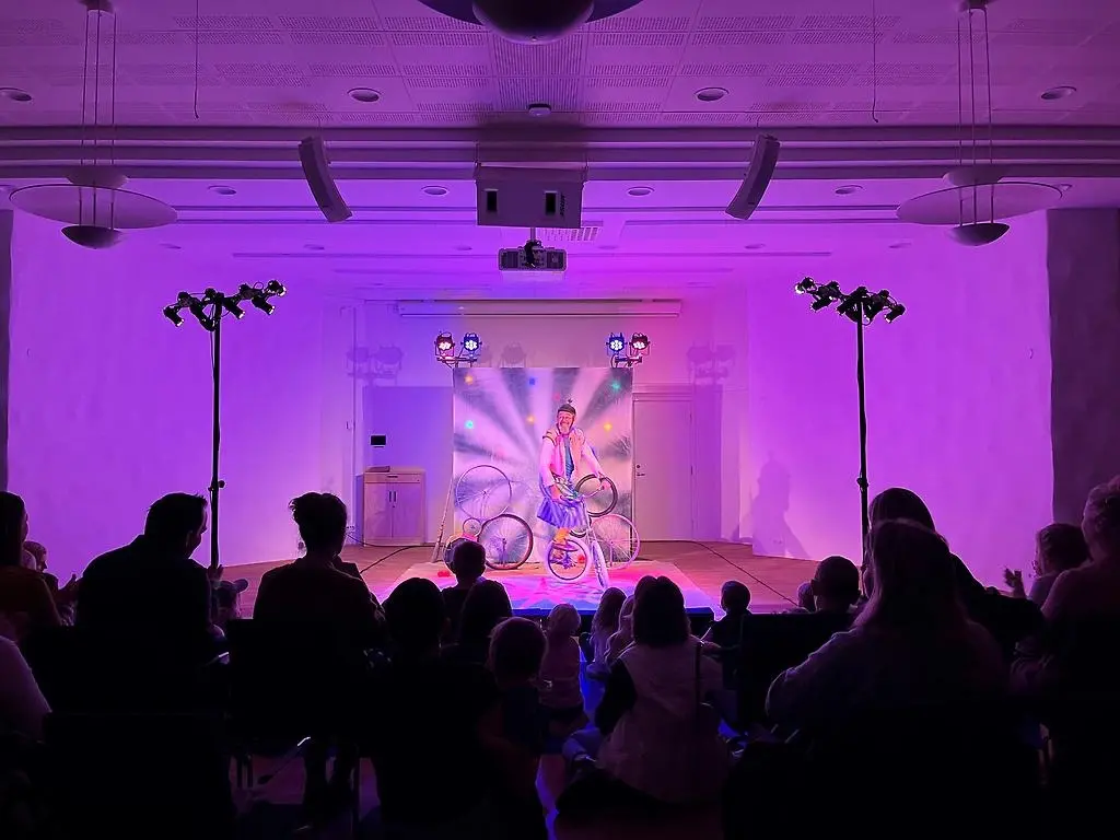 Man som cyklar på en enhjuling framför en publik. Färgglada lampor i bakgrunden och rosa ljus.