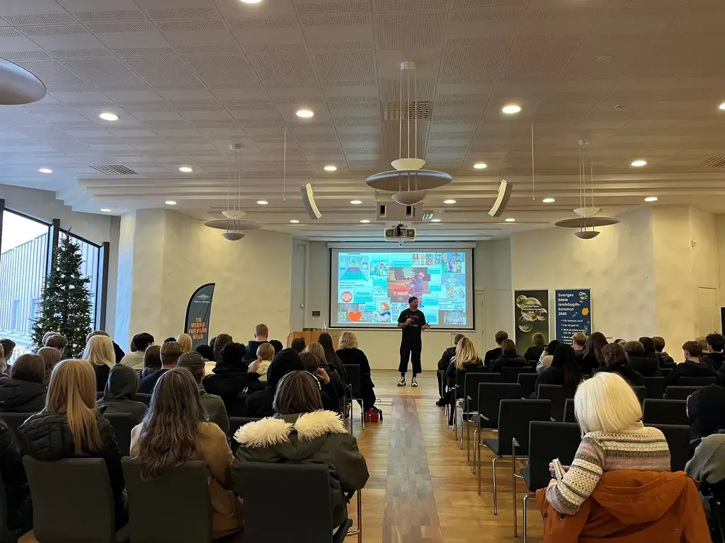 Visar en föreläsningssal där en man står och pratar framför en publik.