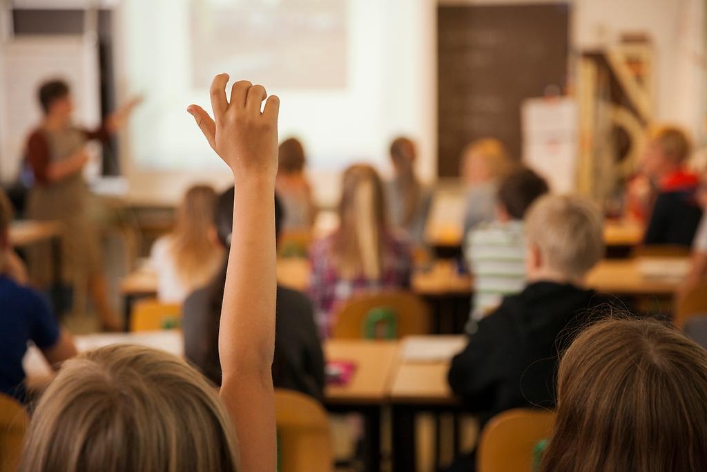Ett barn som räcker upp handen i ett klassrum fullt av elever och en lärare som undervisar. 
