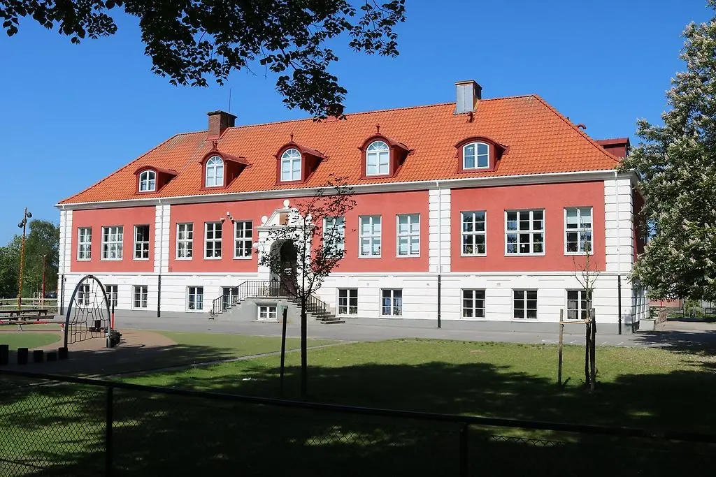 En bild som visar en stor skolbyggnad med röda och vita fasader samt skolgård och gräsmatta.