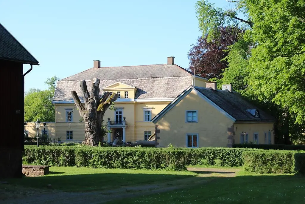 En bild som visar en gul herrgårdsbyggnad, träd, häckar och gräsmatta.