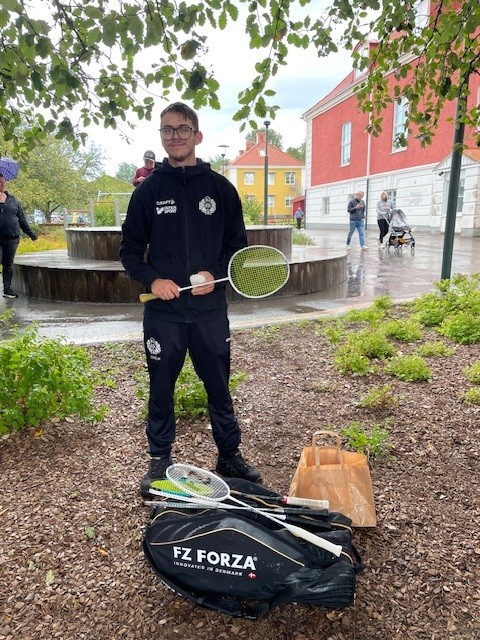 En kortklipt person som står med svarta träningskläder och håller i en vit badmintonsboll samt ett rack.