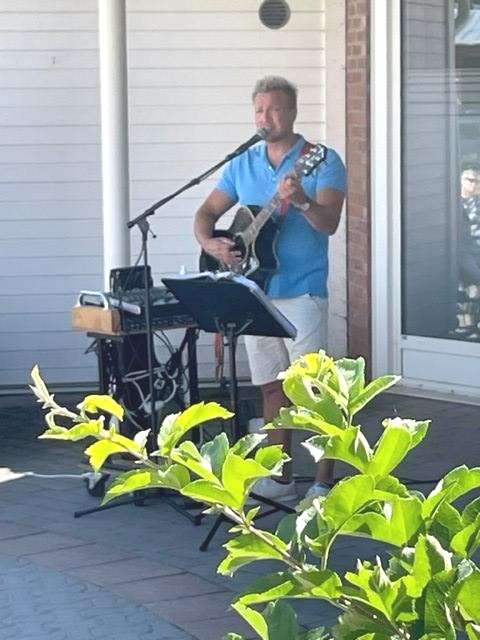 En man i ljusblå tröja står utomhus framför ett mickstativ, sjunger och spelar gitarr.