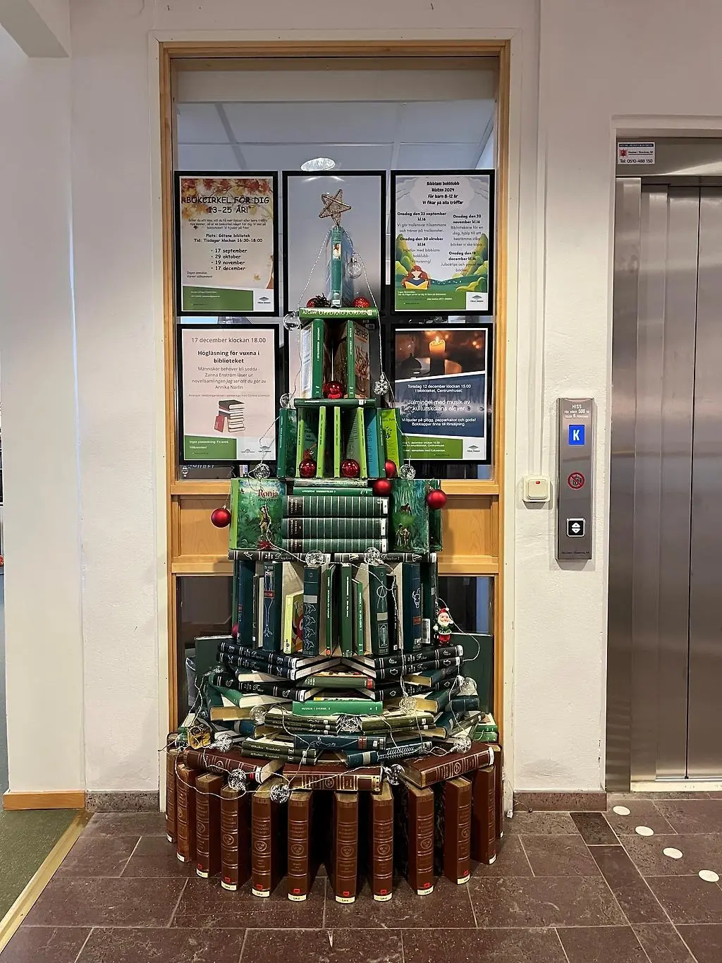 Julgran byggd av olika gröna, blåa och bruna böcker. Pyntad med röda julgranskulor och en ljusslinga samt en guldig stjärna i toppen.