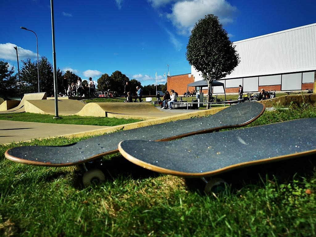 Två stycken svarta skateboards ligger i det gröna gräset. Bakgrunden är en gul skateboardbana.