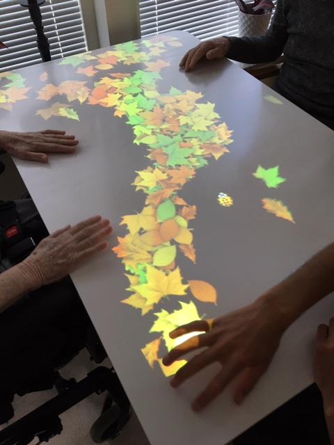 En bild som visar ett av Tovertafelspelen där animerade löv syns på ett bord och händer som rör vid löven. 