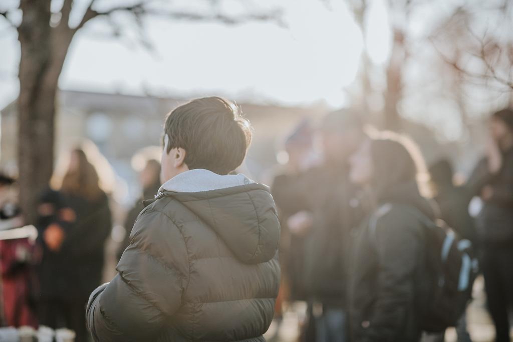 Personer i olika åldrar, står utomhus iklädda vinterkläder. De tittar åt samma håll.