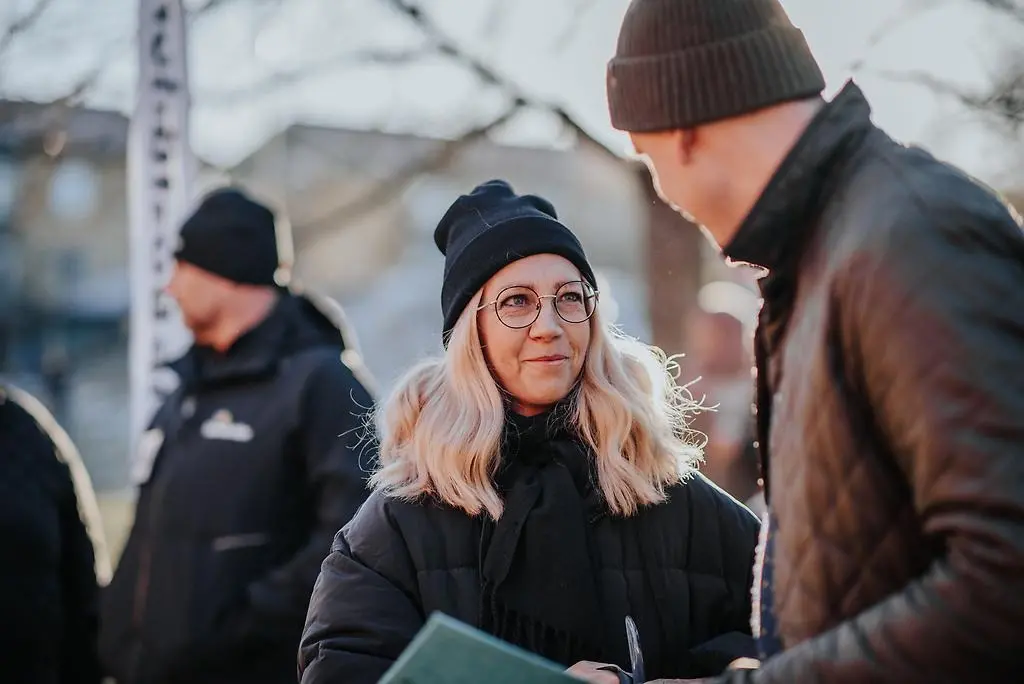 Tre personer som står utomhus varav två syns i profil. En kvinna med blont hår och svarta vinterkläder står i mitten.