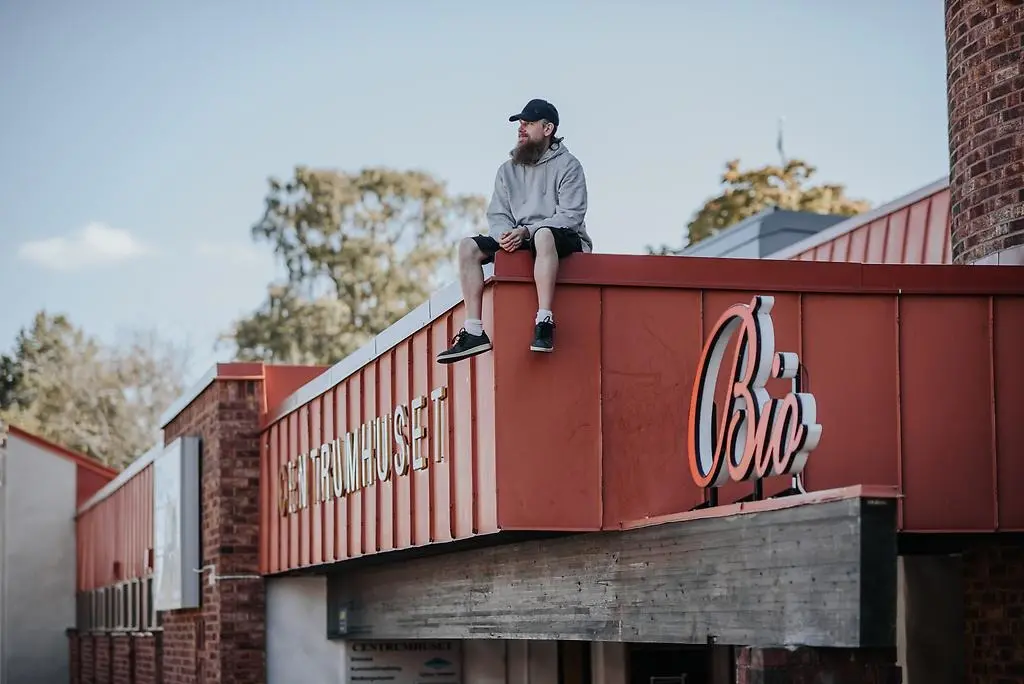 Man som sitter och dinglar med benen på ett tag. Han har shorts, svart keps och en grå luvtröja på sig. 