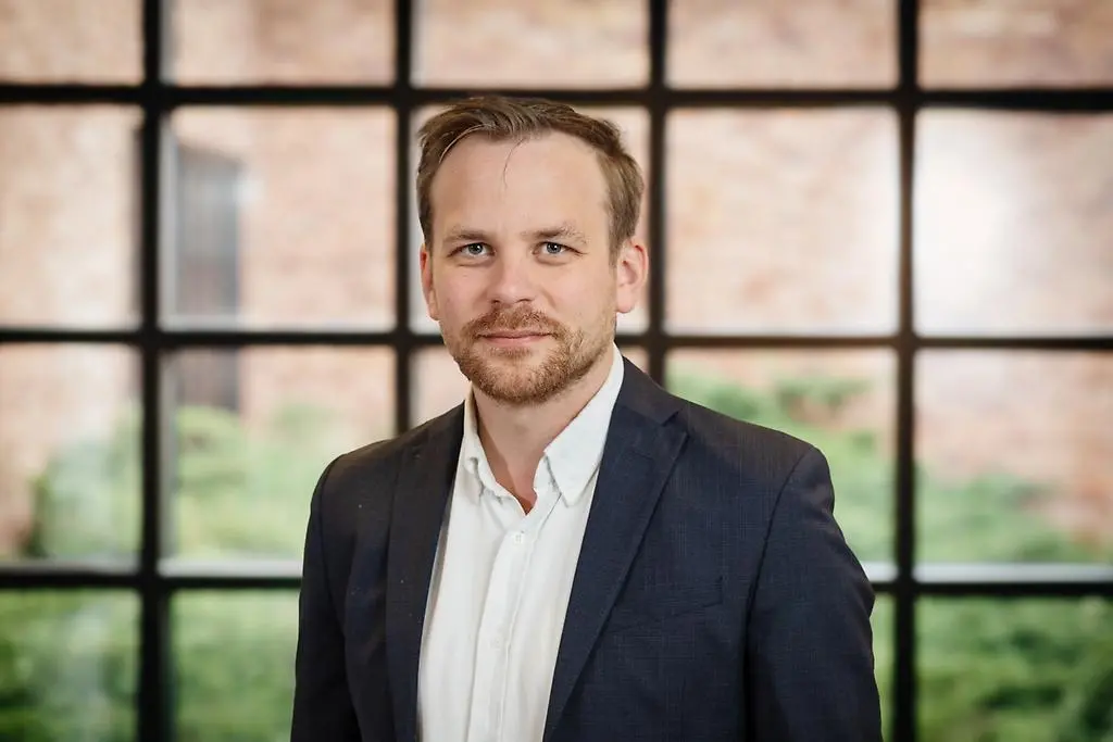 Man som ler och tittar in i kameran. Har en vit skjorta på sig och blå kavaj.