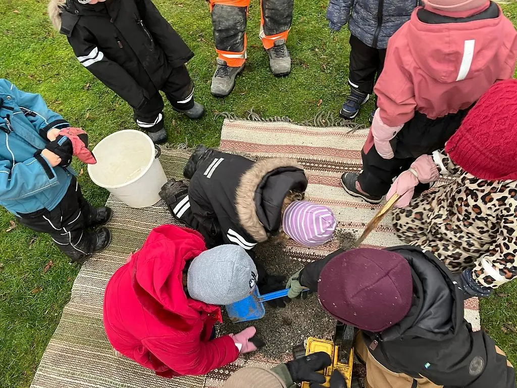 Tre barn jobbar med att gräva en grund till ett kommande lekhus. De sitter på två plastmattor med grus på och har spadar i sina händer. Fyra andra barn och en vuxen ser på. Alla barn har på sig varma vinterkläder.