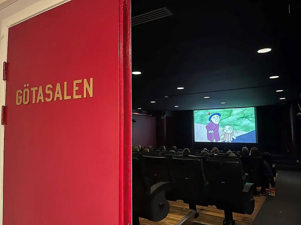 Fotot visar en biosalong bakifrån där publiken sitter och tittar på en bioduk som visar tecknad film med en tant i kappa och hatt och en liten flicka i randig klänning.