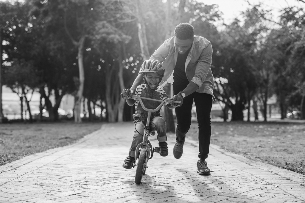 Pojke cyklar och skrattar och man håller i styret och springer bredvid för att hjälpa till att hålla balansen. 