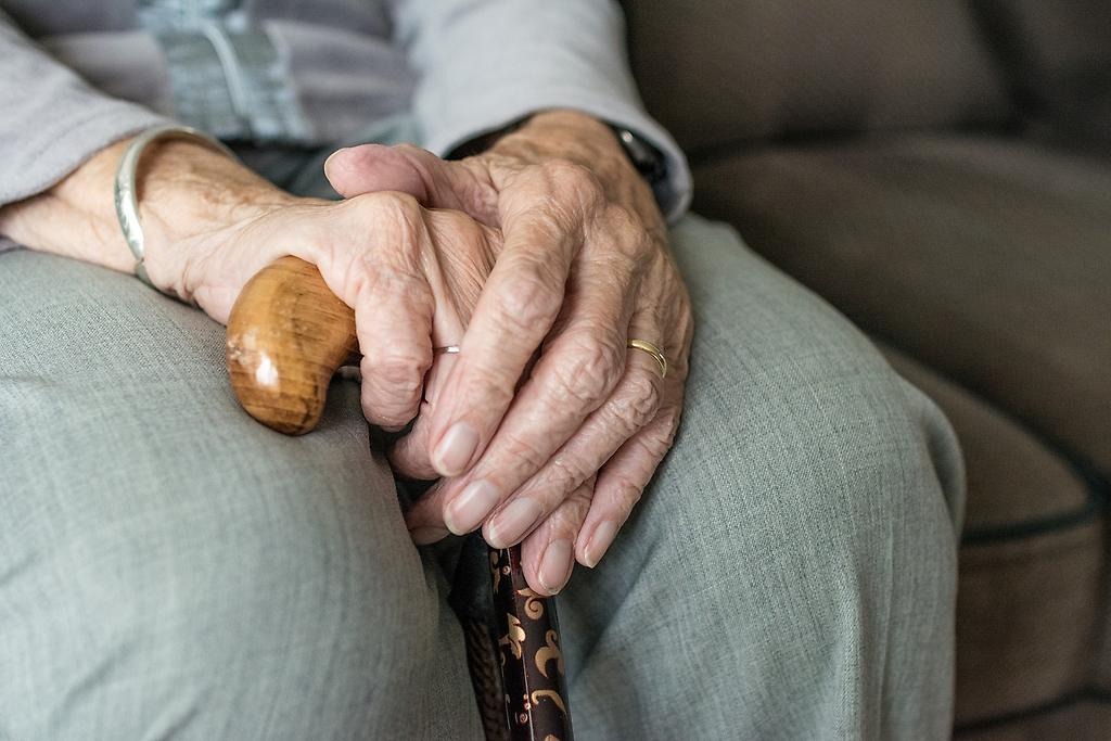 Närbild av en äldre persons händer som håller i en brun träkäpp. Personen har gråa kläder och sitter på en brun soffa. 