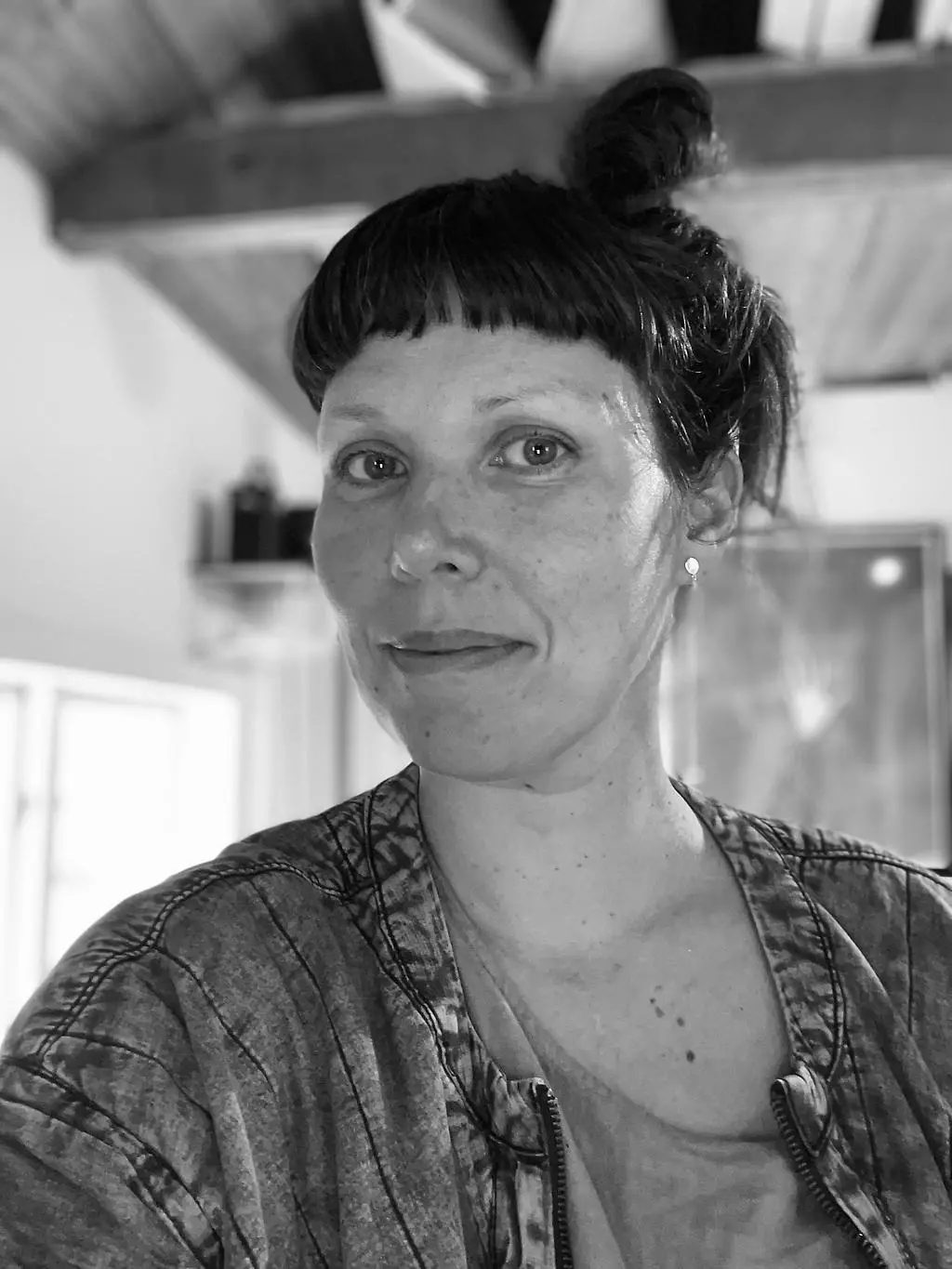 Kvinna med lugg och mörkt hår som är uppsatt i en hög tofs på huvudet. Hon har jeansjacka på sig och ler in i kameran.