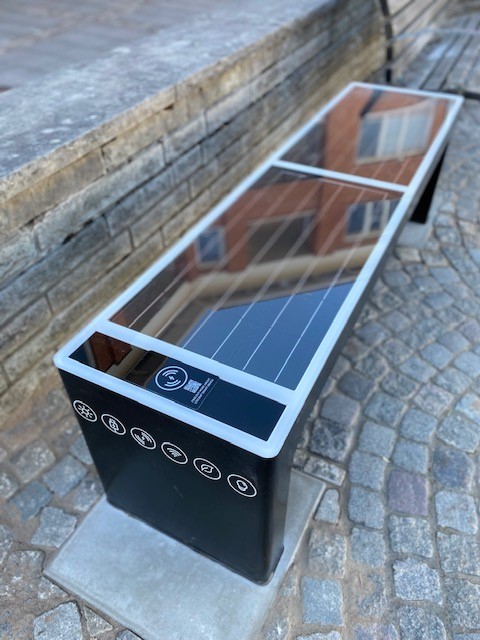 På grå kullersten står en svart bänk med solceller som sittyta. På sidorna sitter laddningsbara uttag för mobilen.