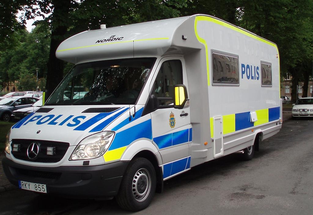 Polisbil i husbilsmodell står parkerad vid en väg. Bilen är vit med blåa och gula reflexmarkeringar. På bilens motorhuv och sida står texten "Polis". 