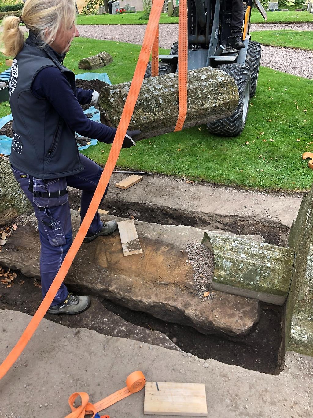 Sten lyftes med hjälp av en maskin från grav med assistans av en kvinna.