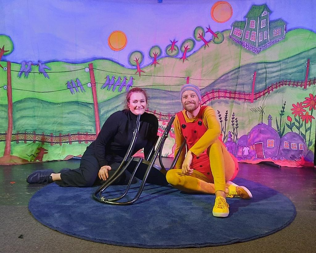 Skådespelarna Jenny och Martin sitter på en rund blå matta framför scendekor som ser ut som ett landskap. Framför sig har de en stol. Jenny har svarta kläder på sig, och Martin har en gul dress med hängsleshorts som är röda med svarta prickar.