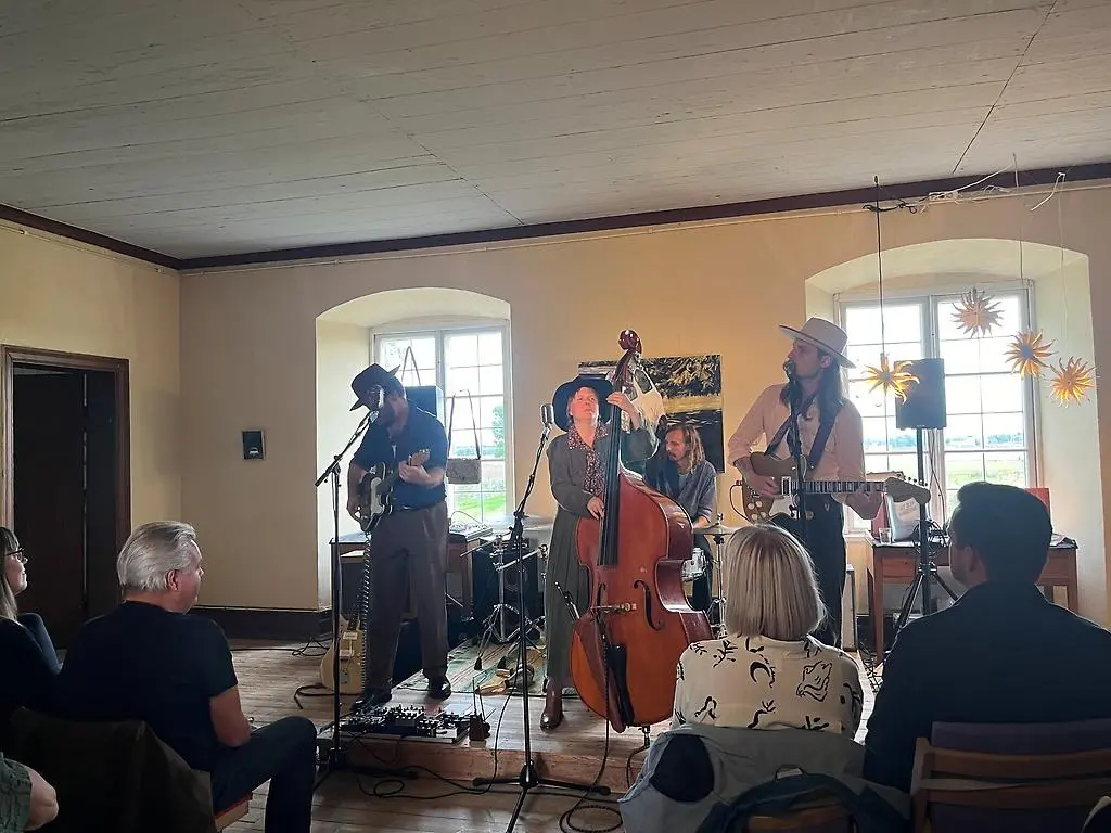 Fyra personer står på en scen med gitarr, trummor och kontrabas. En kvinna och tre män. Alla har hatt på sig.