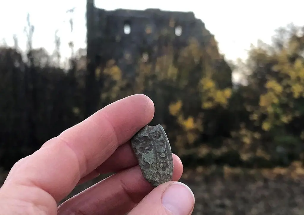 En hand håller upp en liten sten mellan fingrarna med tecken inristade. I bakgrunden syns en gammal stenborg.