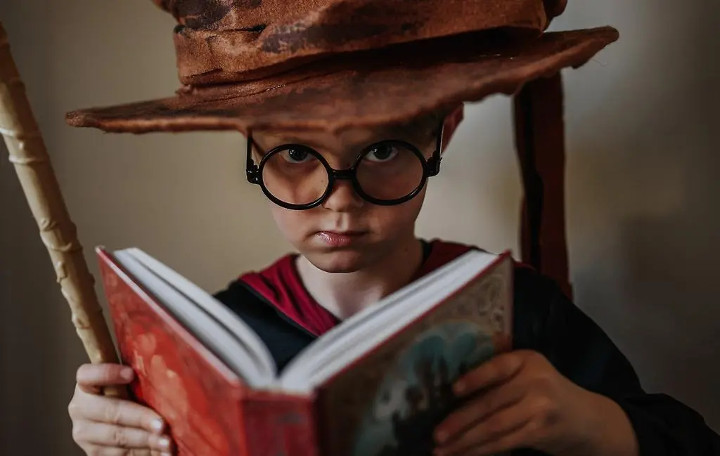 En pojke som har svarta runda glasögon och en brun trollkarlshatt håller i en bok och en trollstav.