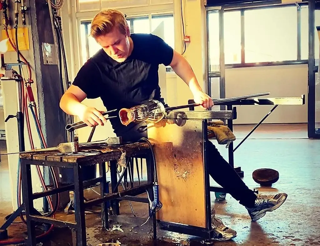 En man sitter vid en arbetsbänk och formar munblåst glas med en tång.
