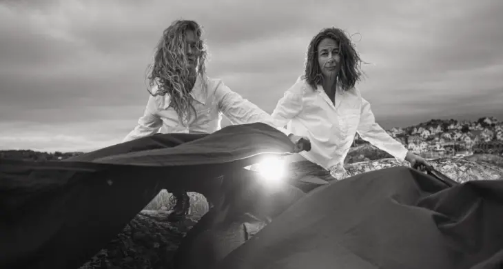 Två kvinnor sitter på huk i vita skjortor med ett svart tyg framför sig som blåser i vinden.