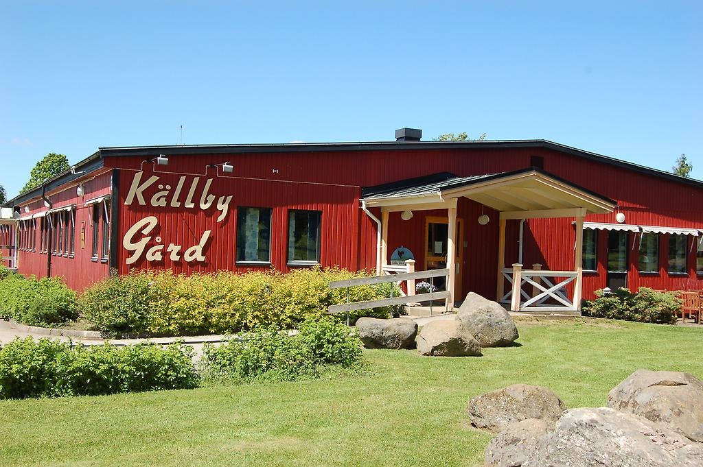 Rött trähus med skylten "Källby gård" på fasaden. Buskar leder fram till entrén.