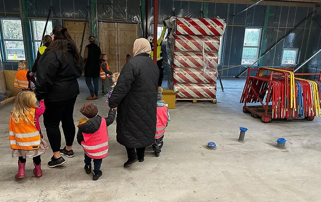 Förskolebarn i reflexvästar tillsammans med sina pedagoger. De tittar på när byggarbetare jobbar i en stor lokal.