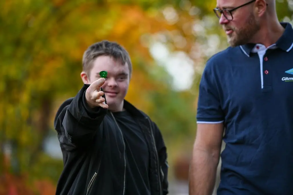 En ungdom med funktionsnedsättning håller upp ett löv och ler. Bredvid går en vuxen.