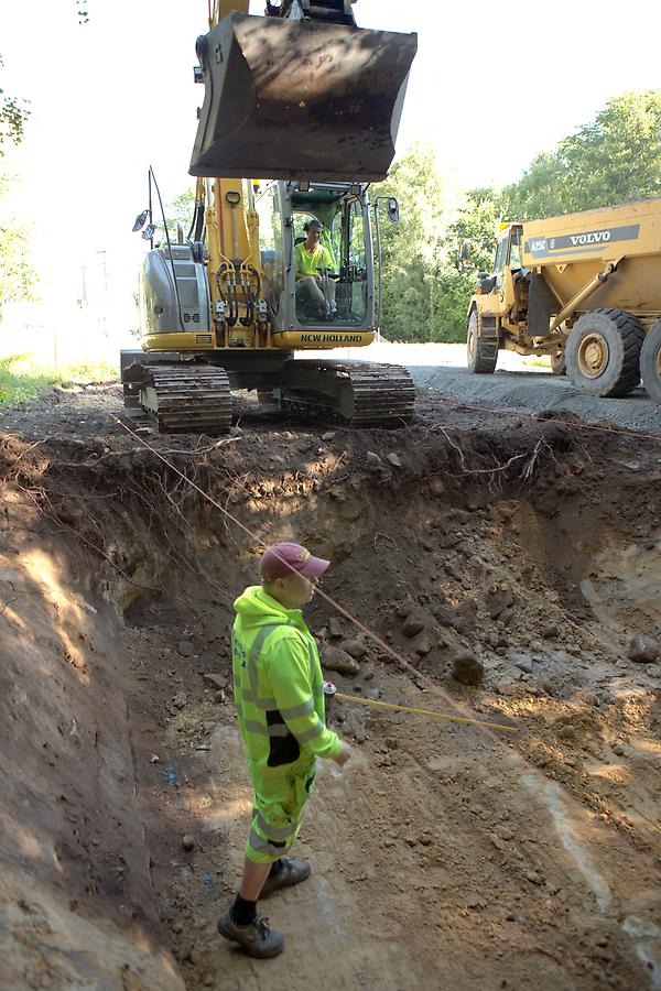En man står i ett grävt hål. Bakom syns grävmaskinen som är redo att fortsätta gräva.
