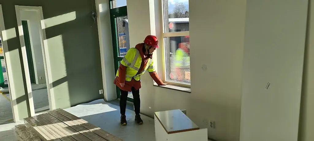 En kvinna iklädd gul reflexväst och en röd hjälm på huvudet, stryker med handen på en fönsterbräda i ett rum med en altandörr och flera stora fönster.