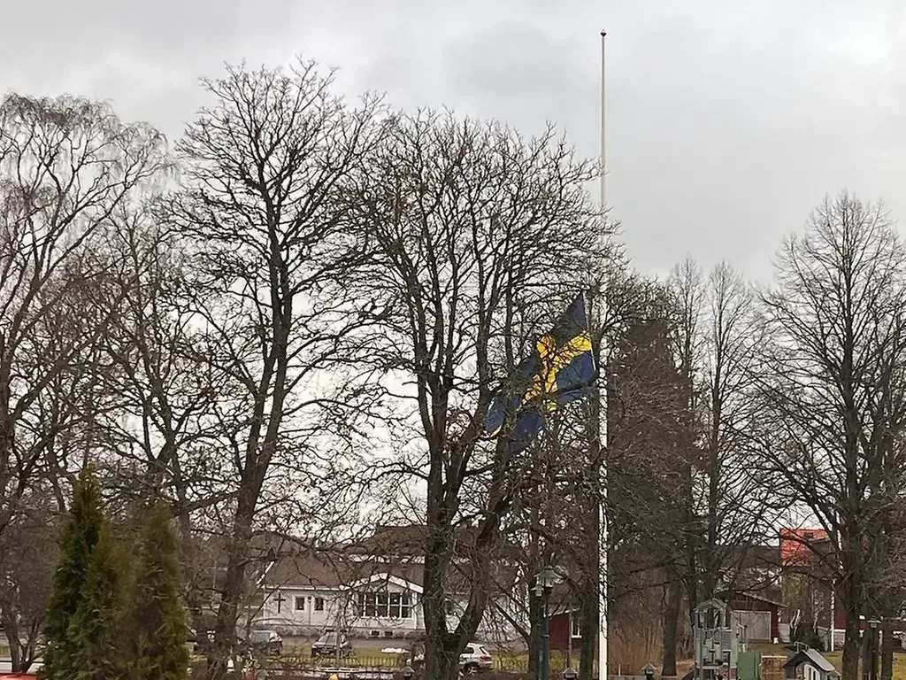 Flagga på halv stång. Träd utan löv och ett beige hus syns i bakgrunden.