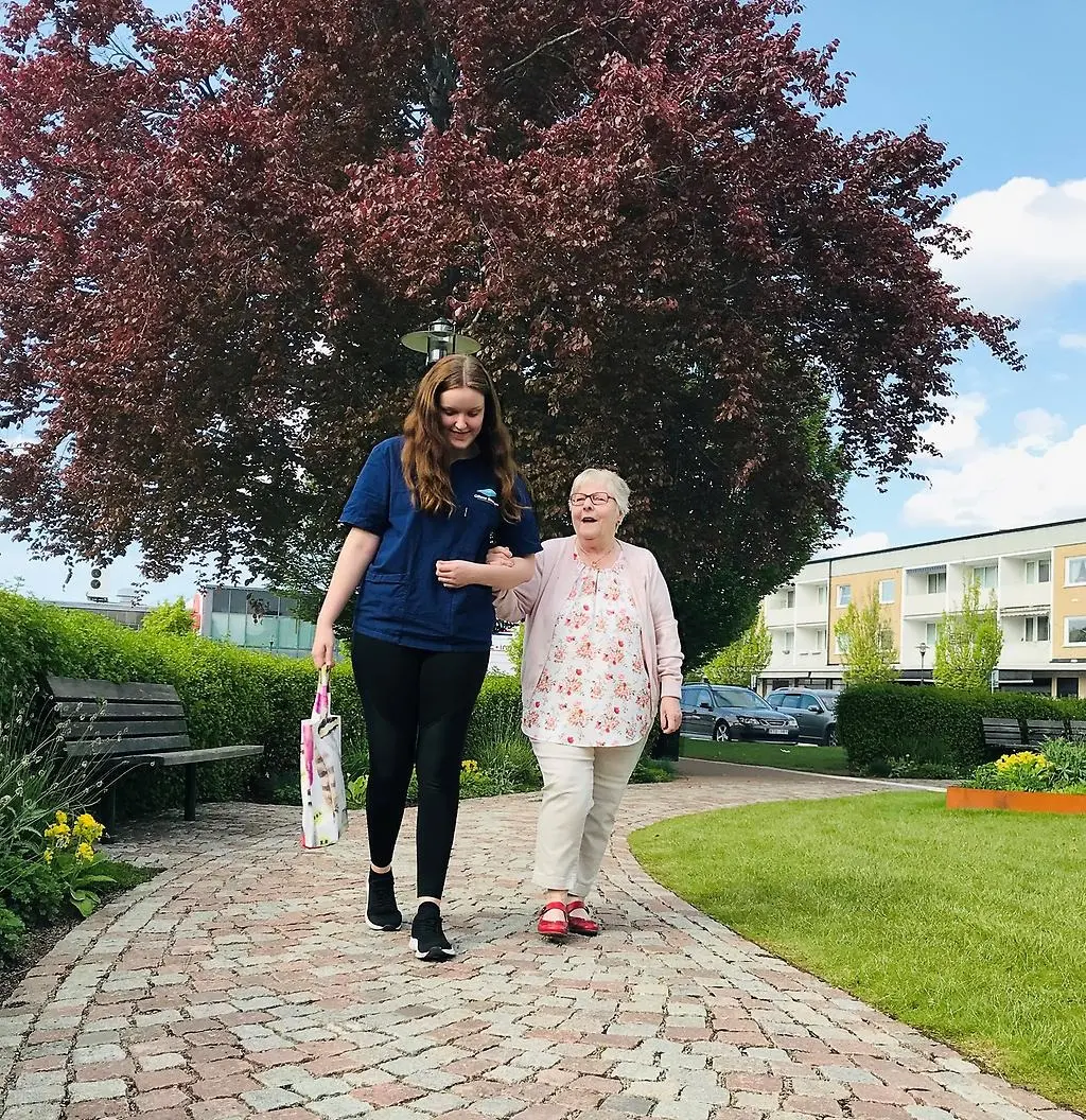 Ung kvinna i vårdklädsel bär en matkasse i ena handen och går i armkrok med en äldre dam.