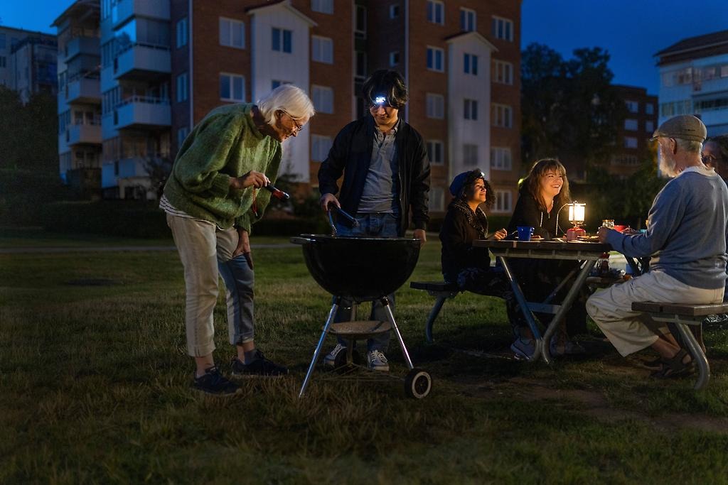 En äldre dam och yngre kille står med en ficklampa vid en grill i mörkret. Bredvid dem sitter personer vid ett bord och äter. 