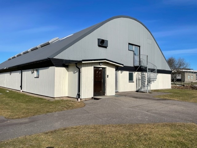 En vit och grå byggnad med runt tak. Bakgrund blå himmel och grönt gräs.