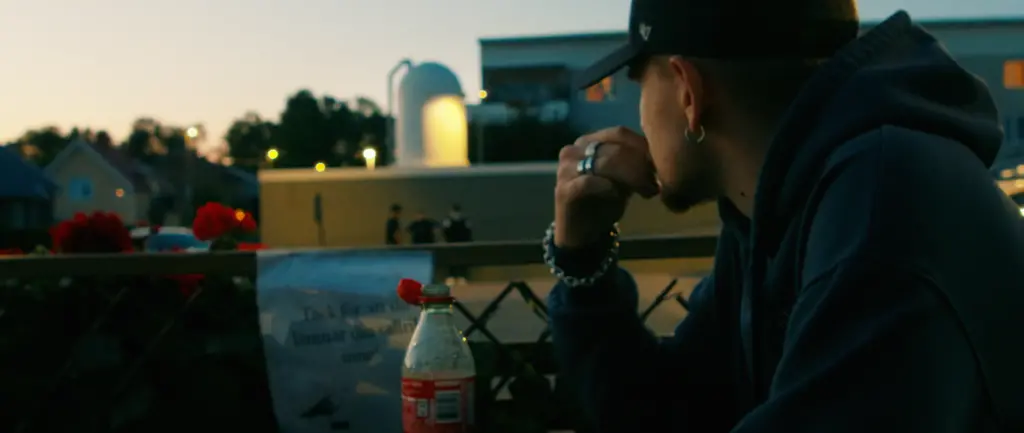 En ung kille i keps sitter ute på en balkong en sommarkväll och tittar bort mot gatan nedanför.