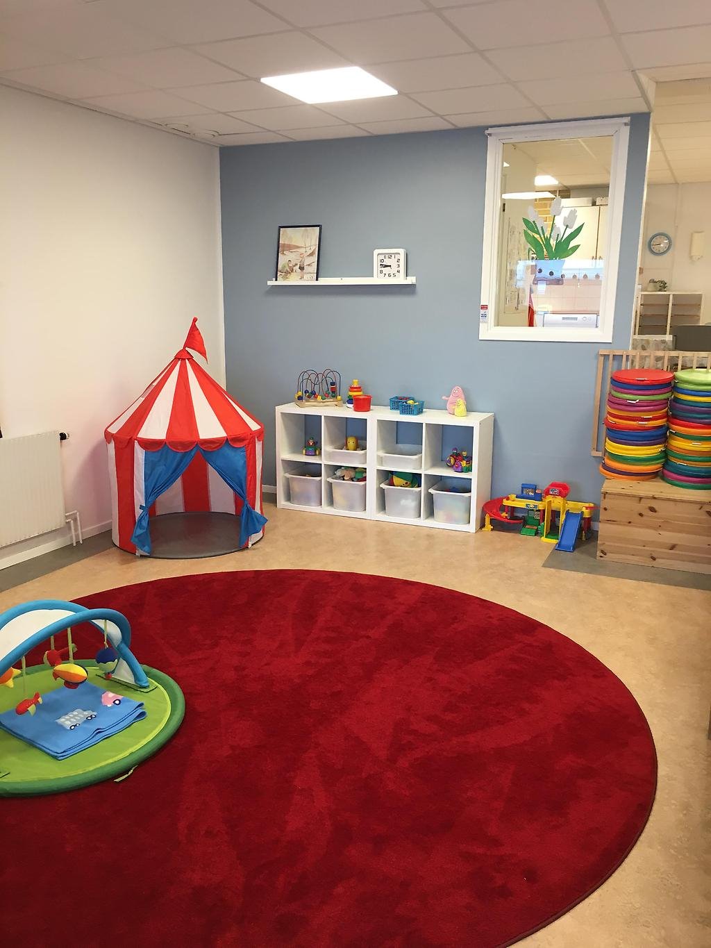 Rum med en stor rund matta i mitten. På mattan står ett babygym, i hörnet finns ett leksakstält och bredvid en hylla fylld med leksaker.