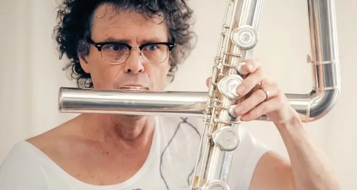 En man med mörkt lockligt hår och glasögon som spelar på en silvrig flöjt.