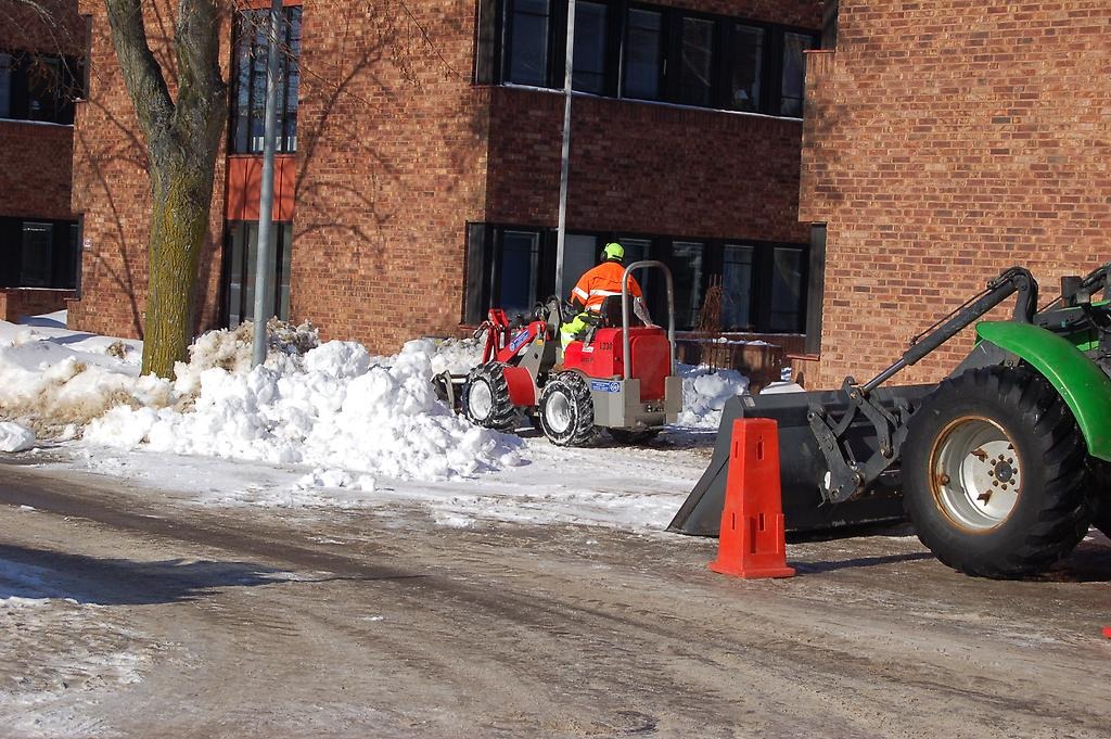 Snöröjare som med plogbil transporterar bort snö från trottoar. 