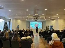 Visar en föreläsningssal där en man står och pratar framför en publik.