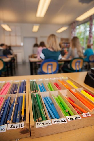 Lådor med färgpennor i förgrunden. I bakgrunden ser man en skolklass som är ur fokus. 