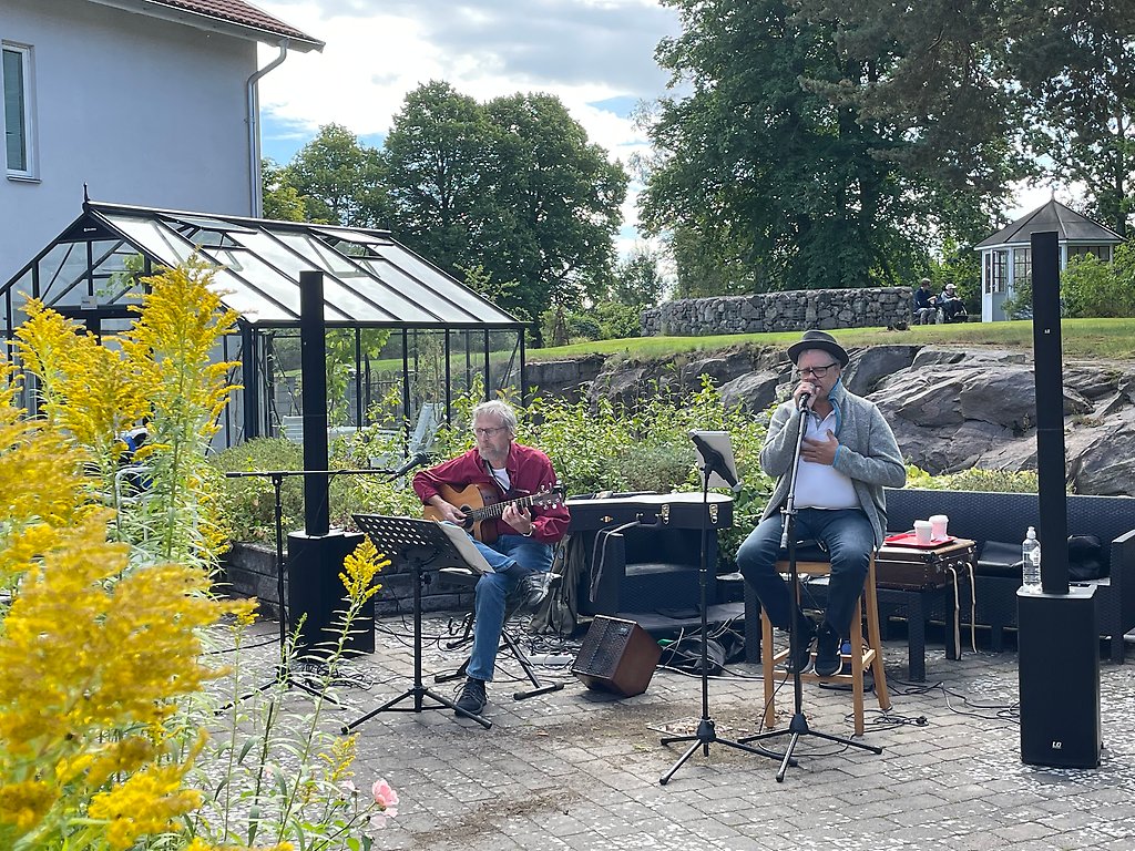 Två män sitter på pallar i trädgården och spelar gitarr och sjunger.