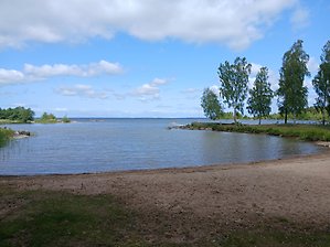 Blombergs badplats. En beige, rund strand med vatten och blå himmel med vita små moln.