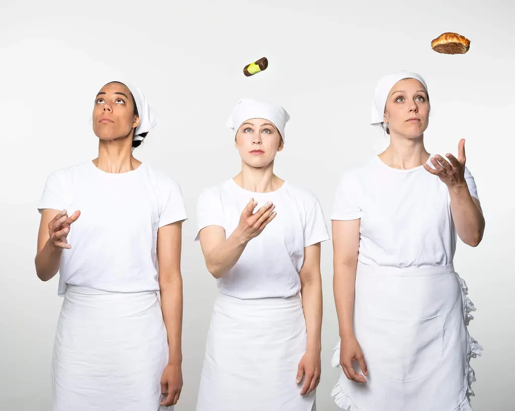 Tre tjejer står i vita bagarkläder och bagarmössor och jonglerar med bakverk.