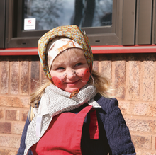 En flicka utklädd till påskgumma med målade kinder och fräknar. Hon har en mönstrad näsduk på huvud och runt halsen. 